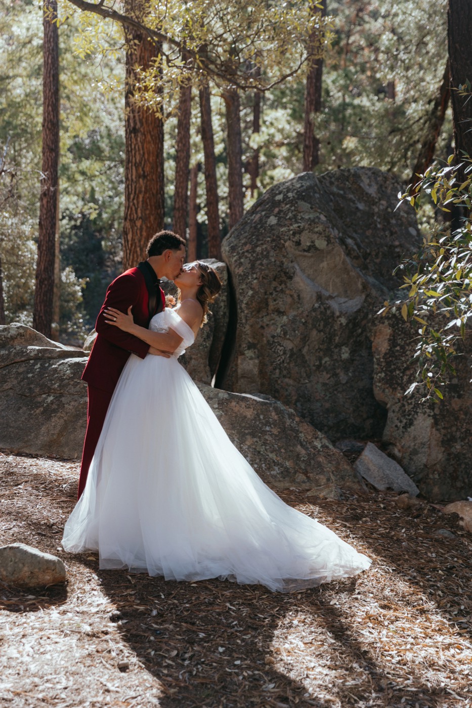 Groom in a deep red suit kissing his bride among pines and granite boulders