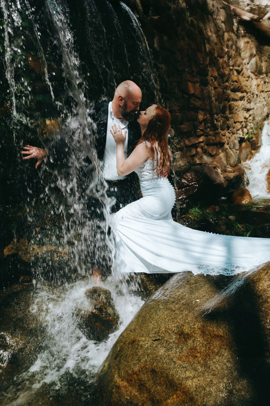 Bride and groom embracing beneath a waterfall, her gown trailing across the rocks